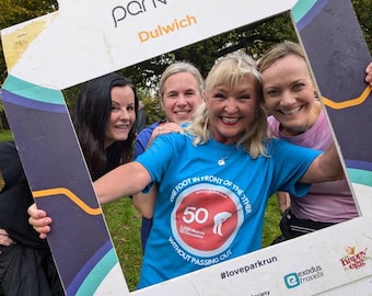 May include: Four women smiling for a photo in a parkrun photo frame. The frame has the text "parkrun Dulwich" and a blue and yellow design. The women are wearing casual clothing and are holding the frame in front of them. The frame also has the text "One foot in front of the other 50 1,000 metres 30 occasions without passing out #loveparkrun".