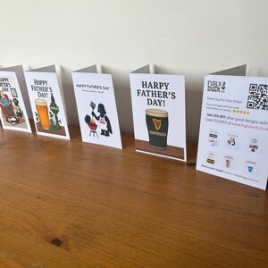 May include: A collection of Father's Day greeting cards displayed on a wooden surface. The cards feature various designs, including beer glasses, a Guinness pint, and humorous illustrations. Text on the cards includes "Happy Father's Day" and "Hoppy Father's Day!"