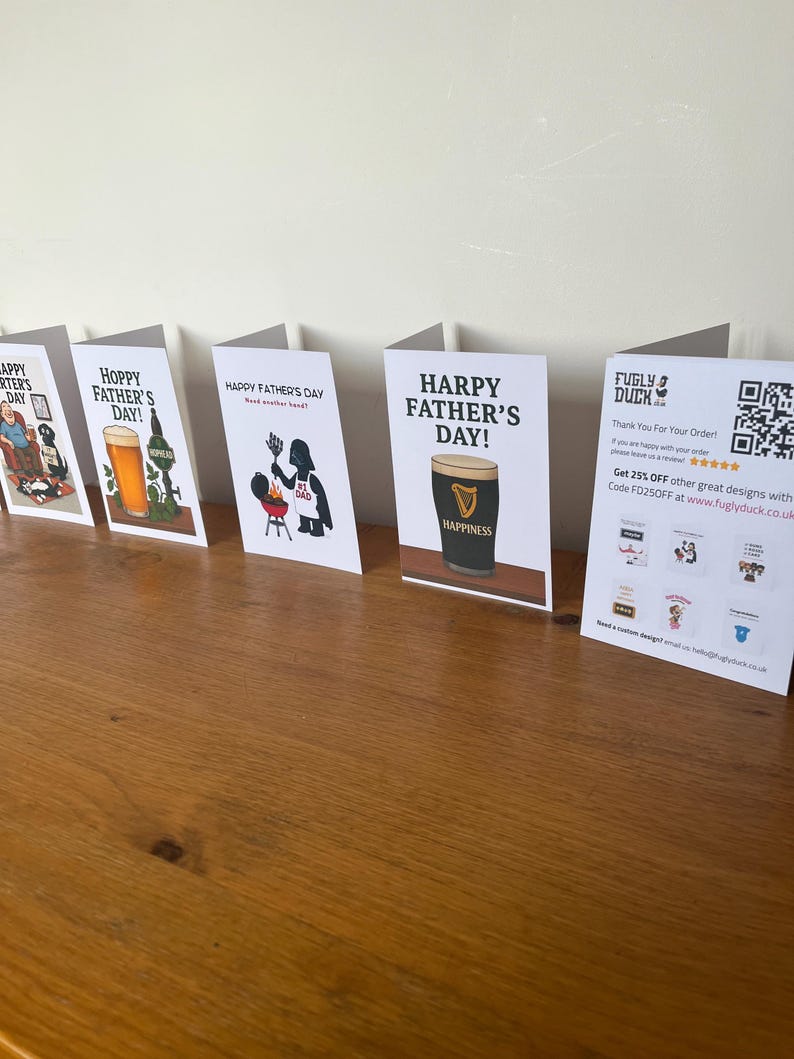 May include: A collection of Father's Day greeting cards displayed on a wooden surface. The cards feature various designs, including beer glasses, a Guinness pint, and humorous illustrations. Text on the cards includes "Happy Father's Day" and "Hoppy Father's Day!"