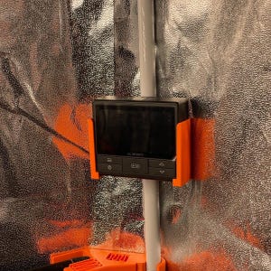 Könnte beinhalten: Ein schwarzer AC Infinity-Controller mit einer orangefarbenen Halterung ist an einer weißen Stange befestigt. Der Controller hat einen Bildschirm und mehrere Tasten. Der Hintergrund ist ein reflektierendes silbernes Material.