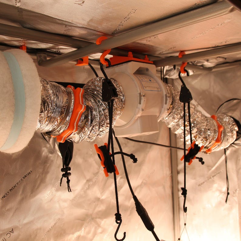 May include: An interior view of a grow tent setup, featuring a white fan, silver ducting, and orange support clips. Black cords with hooks hang from the ceiling, and a white filter is visible on the left. The tent's interior is lined with reflective material.
