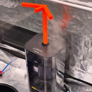 Könnte beinhalten: Ein schwarzer und durchsichtiger Luftbefeuchter mit einem orangefarbenen pfeilförmigen Aufsatz. Der Luftbefeuchter hat eine "MAX"-Anzeige und die Aufschrift "FRONT BOX". Dampf ist sichtbar, und der Hintergrund ist ein reflektierendes silbernes Material.