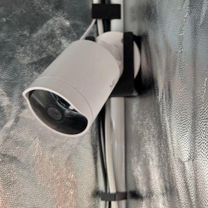 Könnte beinhalten: Eine weiße Überwachungskamera, montiert an einer schwarzen Halterung. Die Kamera hat einen zylindrischen Körper mit dunkler Linse und gebogenem Visier. Kabel sind mit schwarzen Befestigungen an einer reflektierenden silbernen Oberfläche befestigt.