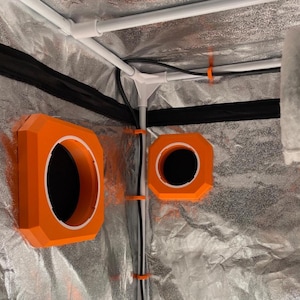 Könnte beinhalten: Innenansicht eines Grow-Zelts mit orangefarbenen Lüftungsöffnungen und einem reflektierenden silbernen Innenfutter. Weiße PVC-Rohre bilden den Rahmen, und schwarze Kabel sind mit orangefarbenen Clips befestigt. Das Zelt ist für den Indoor-Pflanzenanbau konzipiert.