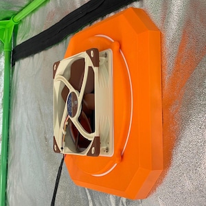 Könnte beinhalten: Eine orangefarbene Lüfterhalterung mit einem weißen und braunen Lüfter. Der Lüfter ist auf einer silbernen, reflektierenden Oberfläche montiert, mit grünen und schwarzen Strukturelementen im Hintergrund. Der Lüfter hat ein schwarzes Netzkabel.