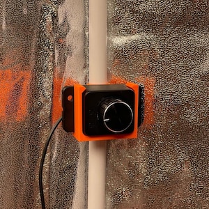 Könnte beinhalten: Ein orangefarbenes und schwarzes Bedienfeld mit einem Drehknopf und einem schwarzen Kabel. Das Panel ist an einer weißen Stange vor einem strukturierten silbernen Hintergrund montiert. Das Zifferblatt hat die Markierungen "ON" und "OFF".