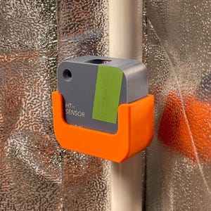 Könnte beinhalten: Ein grauer HT.w-Sensor wird in einer orangefarbenen Halterung gehalten, die an einer weißen Stange befestigt ist. Auf dem Sensor befindet sich ein grüner Aufkleber. Der Hintergrund ist eine strukturierte, metallisch silberne Oberfläche. Der Sensor ist ein kleines, quadratisches Gerät.