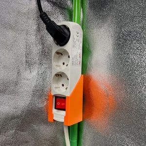 Könnte beinhalten: Eine weiße Steckdosenleiste mit zwei Steckdosen und einem Schalter. Die Steckdosenleiste ist an einer orangefarbenen Wandhalterung befestigt. Die Steckdosenleiste ist an ein grünes Kabel angeschlossen.