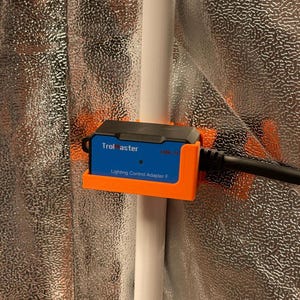 Könnte beinhalten: Ein blauer und schwarzer Lichtsteuerungsadapter mit dem Aufdruck "Trolmaster" und "LMA-14". Der Adapter wird von einer orangefarbenen Halterung gehalten und ist mit einem schwarzen Kabel verbunden. Der Hintergrund ist ein reflektierendes silbernes Material.