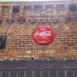 May include: A red Coca-Cola sign with the words "Drink Coca-Cola" on a brick wall. The sign is hanging from chains.