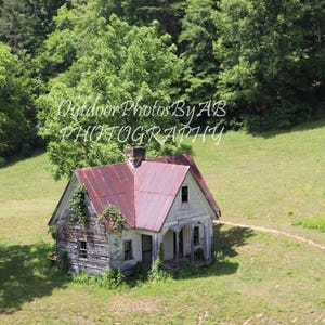 Pode incluir: Uma casa branca e vermelha abandonada com um telhado de metal enferrujado fica em uma encosta gramada. A casa tem uma varanda com uma grade e uma janela no lado.