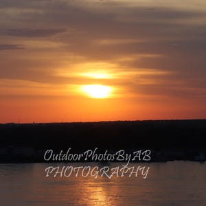May include: A scenic sunset over a body of water with a dark silhouette of trees and land in the foreground. The sky is a vibrant orange and pink with a bright yellow sun in the center. The water is calm and reflective, reflecting the colors of the sky. The text "Outdoor Photos By AB PHOTOGRAPHY" is visible in the lower right corner of the image.