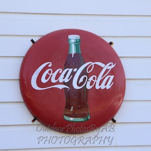 May include: A red, round Coca-Cola sign with a white Coca-Cola logo and a bottle of Coca-Cola in the center.