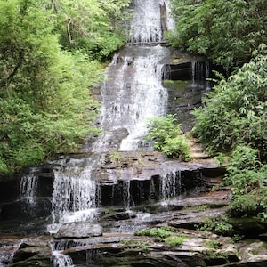 May include: A cascading waterfall flows over layered rock formations, surrounded by lush green foliage. The water plunges into a dark pool below, creating a serene natural landscape. The image is labeled "Outdoor Photos By AB".