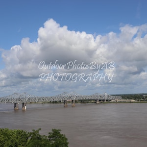 May include: A view of a bridge over a river with a cloudy blue sky. The bridge is made of metal and has a few cars driving on it. The river is wide and calm, and the water is a light brown color. The sky is a light blue with white clouds.