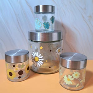 Pode incluir: Um conjunto de quatro frascos de vidro com tampas de aço inoxidável. Os frascos são decorados com desenhos florais e de folhas em amarelo, branco e azul-petróleo. Os frascos variam em tamanho, com o maior no centro e o menor no topo.