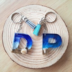 Può includere: Due portachiavi in resina a forma di lettere "D" e "P", con un design a tema spiaggia. Le lettere sono blu con accenti bianchi e color sabbia, e piccole conchiglie. Ogni portachiavi ha una catena e un anello argentati e un nappa.