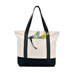 May include: Cream-colored tote bag with black accents. The bag features black straps and a black bottom panel. A front pocket holds green and yellow faux foliage. The bag is designed for carrying items.