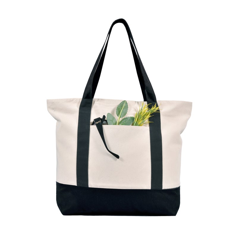 May include: Cream-colored tote bag with black accents. The bag features black straps and a black bottom panel. A front pocket holds green and yellow faux foliage. The bag is designed for carrying items.