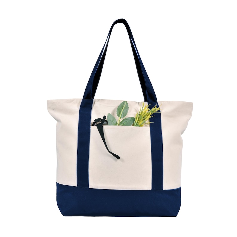 May include: Cream-colored canvas tote bag with navy blue accents. The bag features navy blue handles and a navy blue bottom panel. A front pocket holds green and yellow sprigs. A black strap hangs from the pocket.