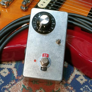 Puede incluir: Un pedal de efectos de guitarra plateado con una perilla negra, un LED rojo y un interruptor de encendido/apagado. El pedal es rectangular y tiene una superficie texturizada. El fondo incluye un cuerpo de guitarra rojo y un cable negro.