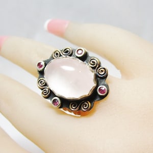 May include: A silver ring with a large oval rose quartz stone set in a filigree design. The ring is accented with small pink gemstones.