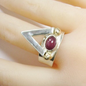 May include: A silver ring with a triangular design and a red gemstone. The ring is set with small gold beads.