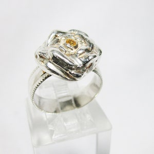 May include: A silver ring featuring a detailed rose design with a small, yellow gemstone at the center. The ring has a textured band and is displayed on a clear stand against a white background. The floral design is the focal point.