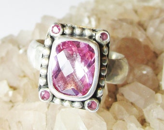 May include: A silver ring with a pink gemstone set in a square bezel. The ring is surrounded by small silver beads and has four smaller pink gemstones on the corners.