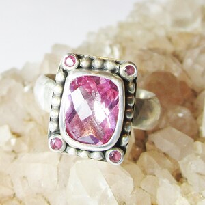 Könnte beinhalten: Ein silberner Ring mit einem rosafarbenen Edelstein, der in einer quadratischen Fassung sitzt. Der Ring ist mit kleinen silbernen Perlen verziert und hat an den Ecken vier kleinere rosafarbene Edelsteine.