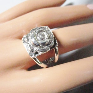 May include: A silver ring with a raised rose design. The ring is worn on a finger.