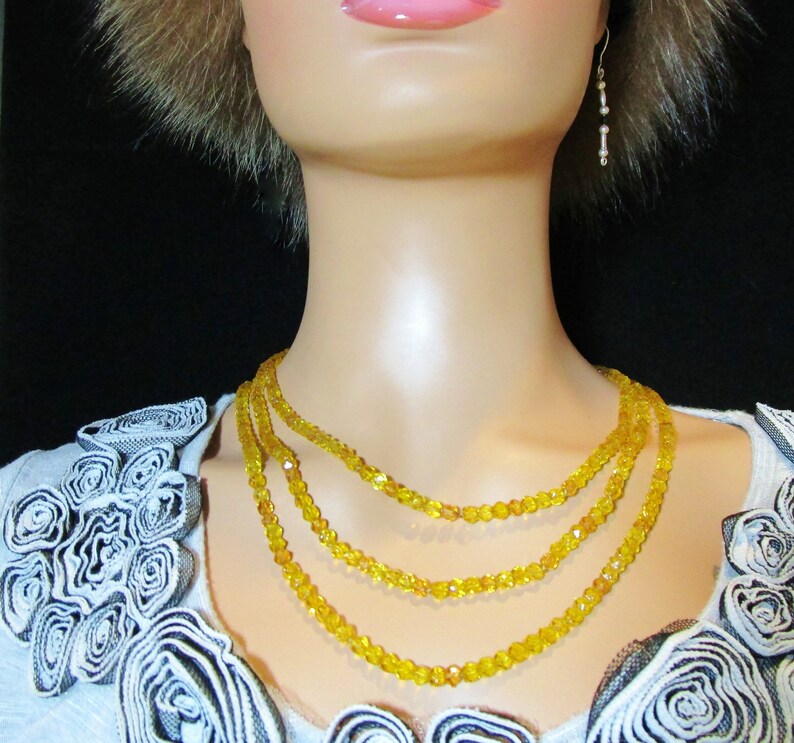Puede incluir: Un collar de tres hilos con peque&ntilde;as cuentas amarillas facetadas. El collar est&aacute; en capas y descansa sobre el cuello de un maniqu&iacute;. Tambi&eacute;n se ven un par de pendientes colgantes con peque&ntilde;as cuentas.
