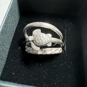 May include: A set of three stacked silver-toned rings resting inside a black box. The top ring has a textured finish, the middle ring features a three-dimensional sea turtle charm, and the bottom ring displays a continuous braided or twisted pattern.