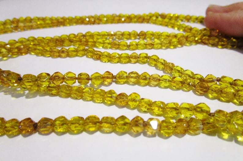 Puede incluir: Primer plano de varios hilos de cuentas amarillas facetadas. Las cuentas est&aacute;n ensartadas, creando un collar o elemento decorativo. Las cuentas tienen una superficie brillante y reflectante, y el color es un amarillo dorado c&aacute;lido.