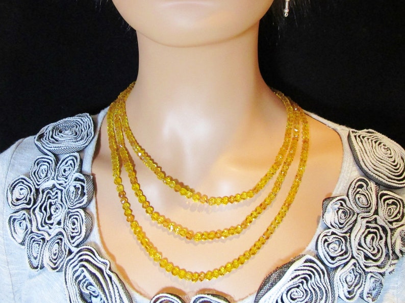 Puede incluir: Un collar de tres hilos con peque&ntilde;as cuentas amarillas facetadas. El collar es en capas y se usa en un maniqu&iacute;. El maniqu&iacute; lleva una camiseta gris claro con rosetas decorativas.