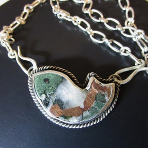 May include: A silver necklace with a unique pendant featuring a green, white, and copper stone set in a silver frame. The pendant is shaped like a bird.