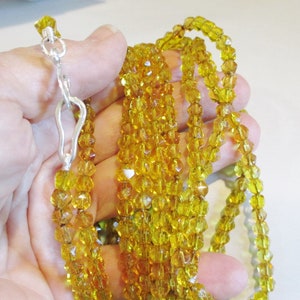 Puede incluir: Un collar de m&uacute;ltiples hilos con peque&ntilde;as cuentas facetadas de color &aacute;mbar. El collar se sujeta con un cierre plateado. Las cuentas tienen una calidad brillante y transl&uacute;cida, y el collar se muestra sobre un fondo neutro.