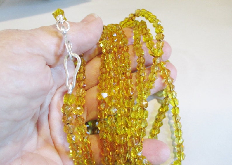 Puede incluir: Un collar de m&uacute;ltiples hilos con peque&ntilde;as cuentas facetadas de color &aacute;mbar. El collar se sujeta con un cierre plateado. Las cuentas tienen una calidad brillante y transl&uacute;cida, y el collar se muestra sobre un fondo neutro.