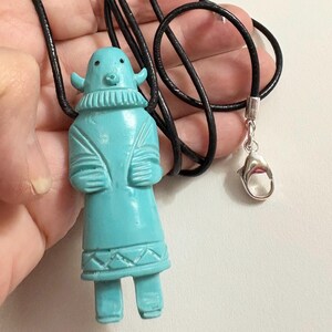 May include: A turquoise-colored pendant necklace featuring a carved figure with a detailed design. The figure has a stylized face, arms, and legs. The pendant hangs from a black cord with a silver-tone clasp. The necklace is a unique piece of jewelry.