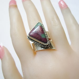 May include: A silver ring with a triangular green and pink gemstone set in a silver bezel. The ring band is wide and has a textured finish.