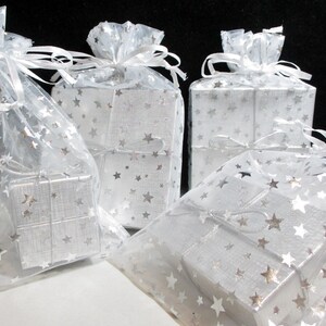Puede incluir: Varias cajas de regalo plateadas peque&ntilde;as, envueltas en papel con estampado de estrellas y encerradas en bolsas transparentes tambi&eacute;n decoradas con estrellas. Cada bolsa est&aacute; atada con una cinta blanca. Los regalos est&aacute;n sobre una superficie blanca sobre un fondo negro.