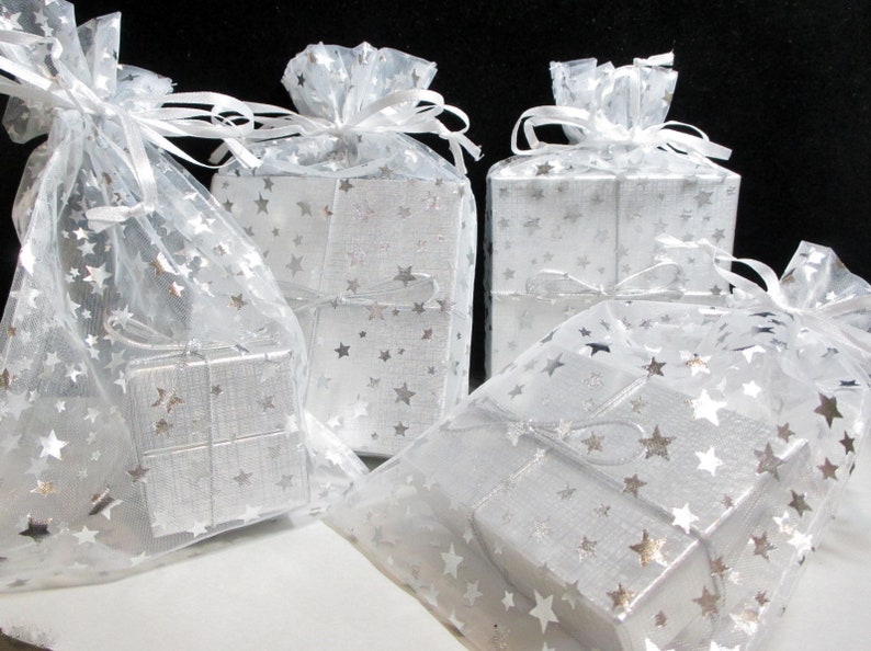 Puede incluir: Varias cajas de regalo plateadas peque&ntilde;as, envueltas en papel con estampado de estrellas y encerradas en bolsas transparentes tambi&eacute;n decoradas con estrellas. Cada bolsa est&aacute; atada con una cinta blanca. Los regalos est&aacute;n sobre una superficie blanca sobre un fondo negro.