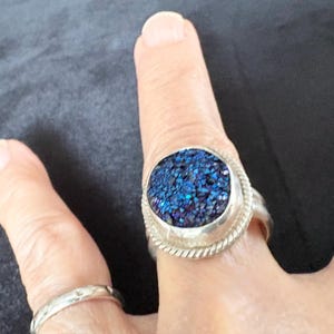 May include: A silver ring featuring a large, circular, deep blue druzy stone with a textured, crystalline surface. The stone is set in a polished silver bezel surrounded by a decorative twisted rope border. A plain silver band is also visible.