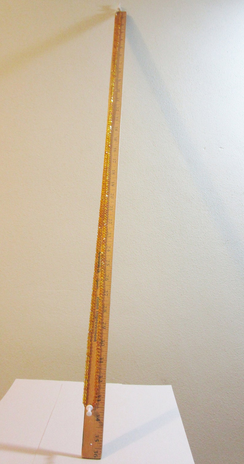 Puede incluir: Una regla de madera, de aproximadamente 91 cm de largo, con marcas en pulgadas y n&uacute;meros. Un hilo de cuentas de color &aacute;mbar est&aacute; drapeado a lo largo de la regla. La regla est&aacute; en &aacute;ngulo contra una superficie blanca y una pared beige.