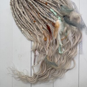 Op de afbeelding: Handgemaakte dreadlocks in grijstinten en blond, versierd met veren, kralen en decoratieve wikkelingen. De dreadlocks hebben een golvende, getextureerde uitstraling en zijn op een wit oppervlak geplaatst.