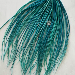 May include: A bundle of teal and turquoise dreadlocks with silver decorative charms. The dreadlocks are a mix of straight and wavy textures, creating a layered effect. The hair extensions are arranged on a white wooden surface.