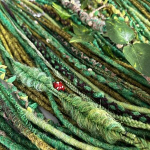 Può includere: Una collezione di dreadlock verdi e marroni, adornati con foglie finte e un piccolo fungo rosso. I dreadlock variano nelle tonalità di verde, creando un'estetica naturale e terrosa. Alcuni dreadlock sono intrecciati o avvolti con elementi decorativi.