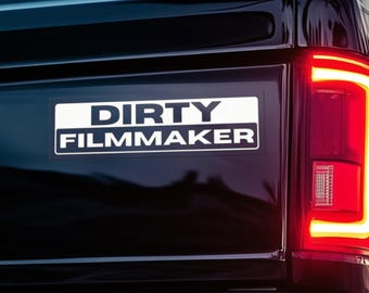 May include: A rectangular white and black bumper sticker with the words "DIRTY FILMMAKER" in bold, sans-serif font. The sticker is affixed to the black rear of a vehicle, near a red taillight.