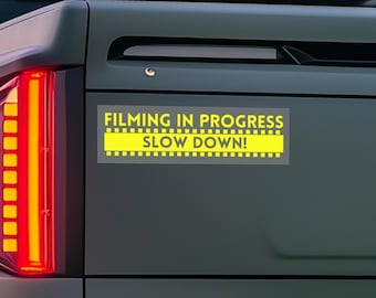 May include: A yellow and black bumper sticker with the text "Filming in Progress Slow Down!" on a gray truck.
