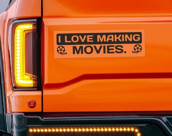 May include: Orange pickup truck with a black bumper and a sticker that reads "I LOVE MAKING MOVIES." with film reel icons on either side of the text.